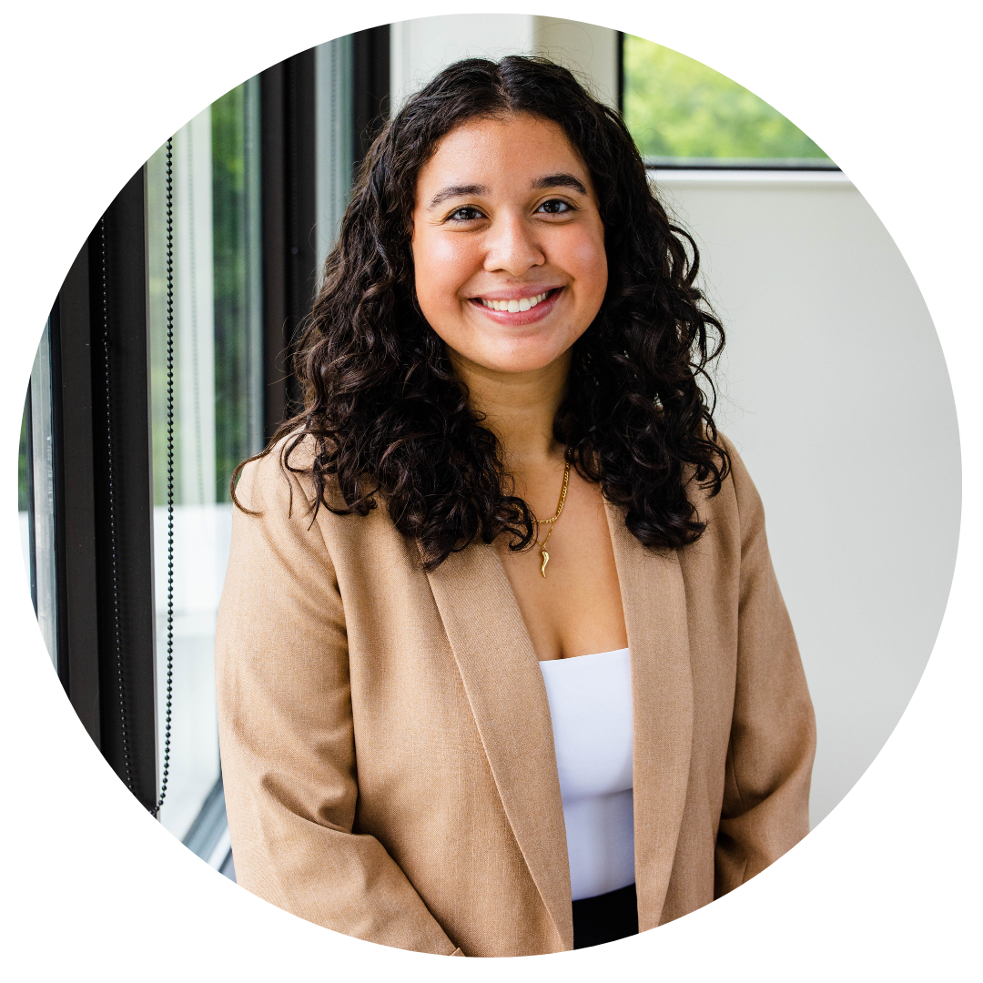 A woman with long curly hair, wearing a tan blazer and white top, smiles while standing indoors near large windows.