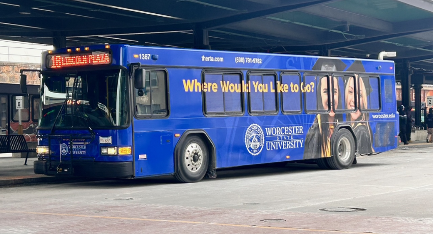A blue city bus wrapped in a Worcester State University advertisement is parked at a transit terminal, proudly displaying the university's slogan, "Where Would You Like to Go?" on its side.