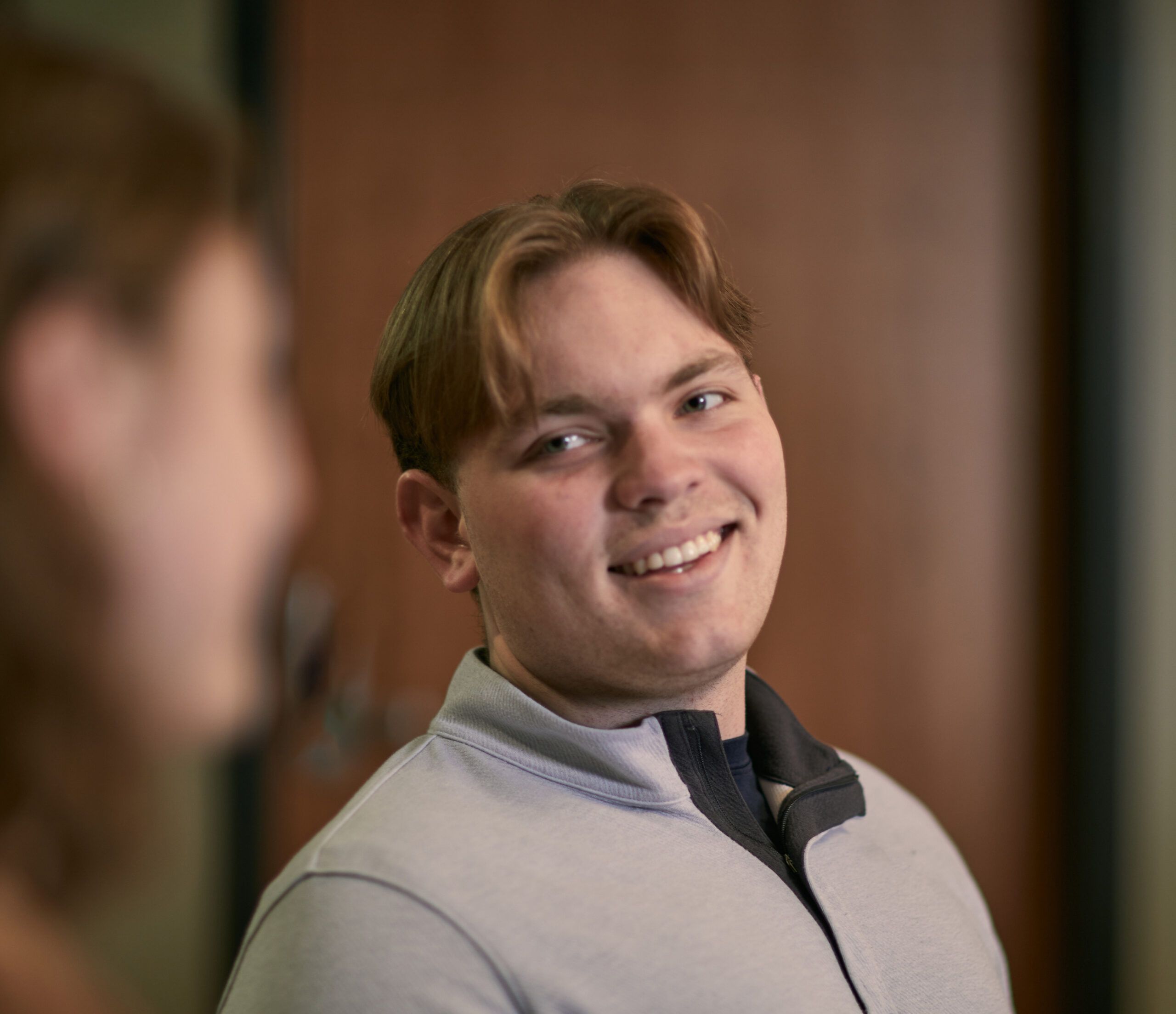 A young man with light brown hair and a light gray zip-up shirt smiles while sitting indoors, possibly discussing tuition and financial aid, with another person blurred in the foreground.