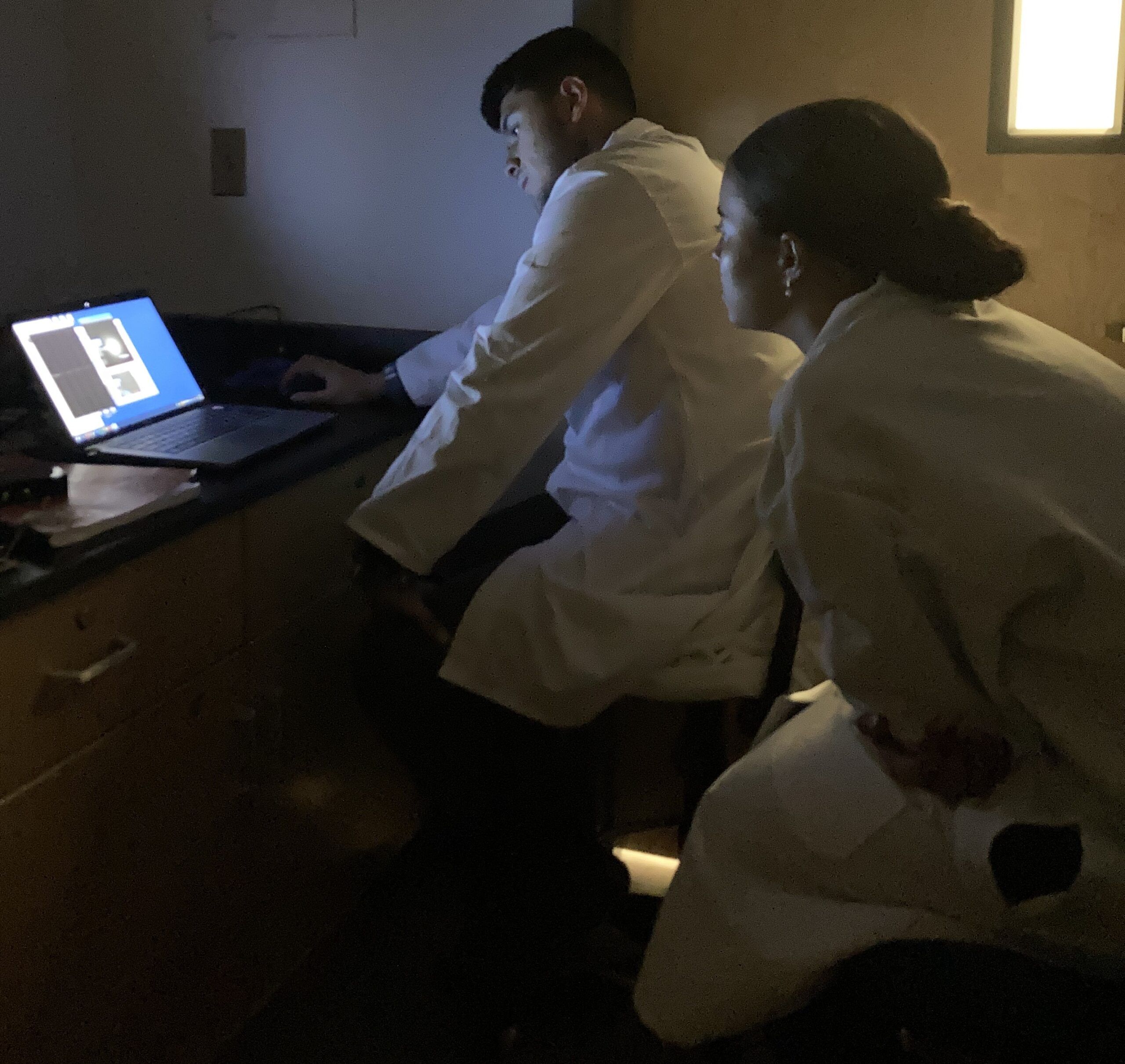 Two people in lab coats sit in a dimly lit room, looking at medical images on a laptop screen.