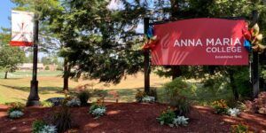 Large red sign reads "Anna Maria College, Established 1946" in front of a landscaped garden with trees and a banner on a pole.