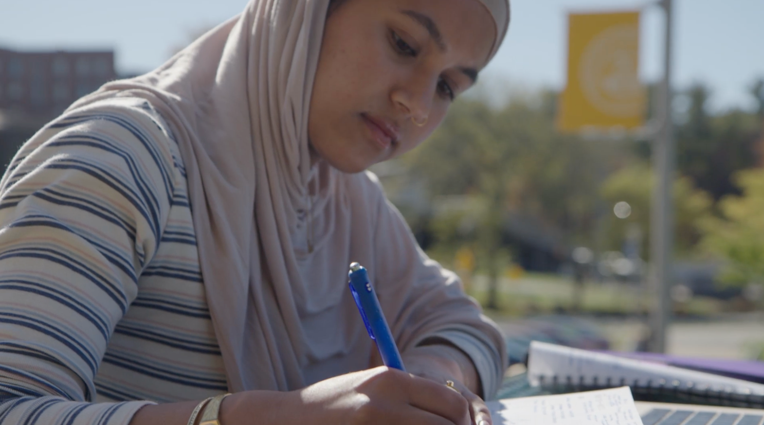 About: A person wearing a light-colored hijab and striped shirt writes with a blue pen in a notebook outdoors, with a blurred campus background.
