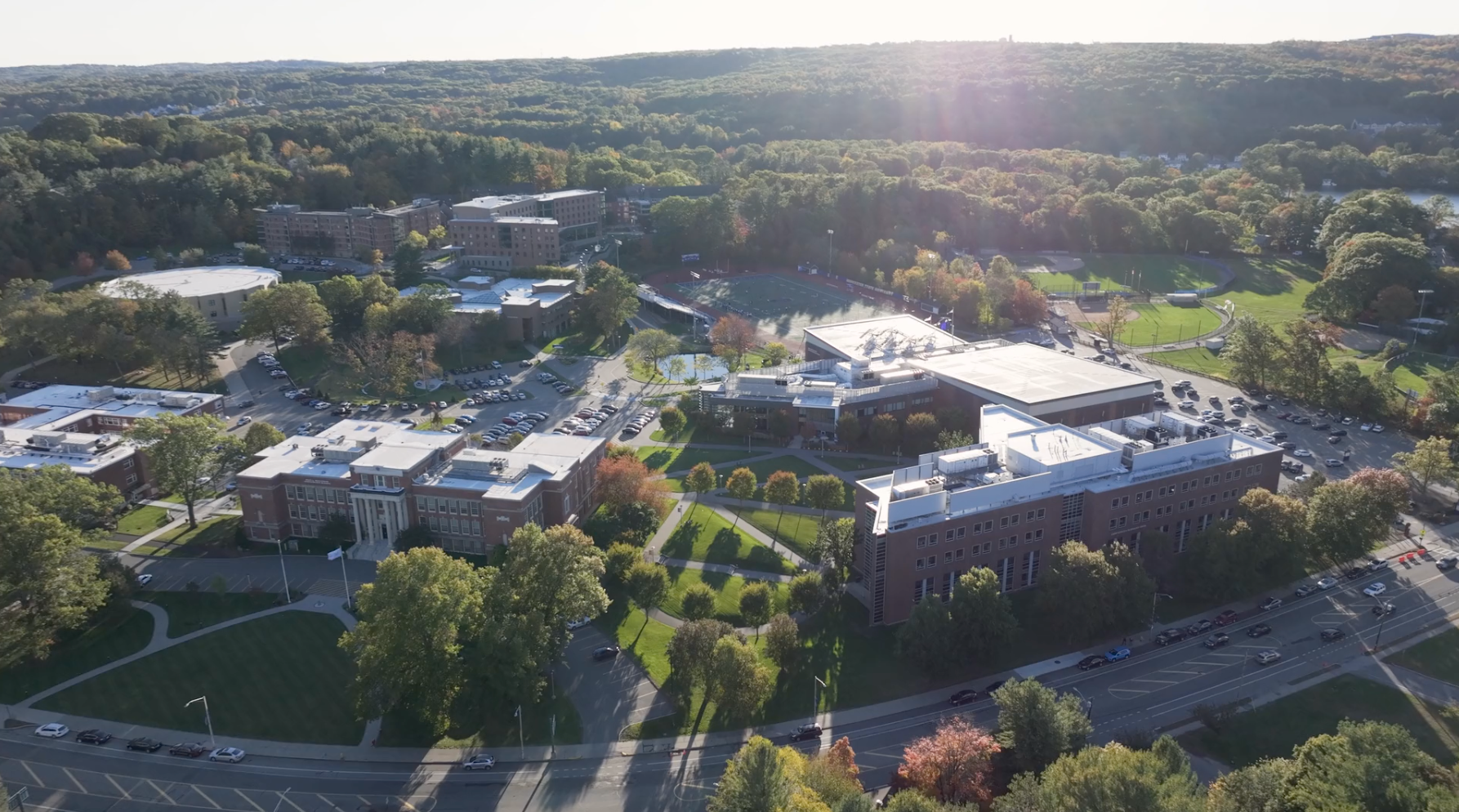 Aerial view of a college campus with multiple buildings, parking lots, sports fields, and surrounding trees under sunlight, where students explore opportunities for tuition and financial aid.