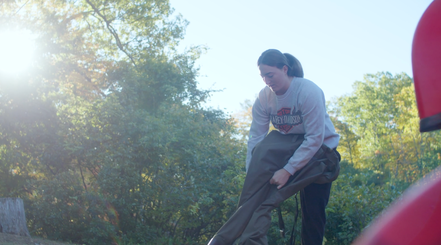 A person outdoors in daylight puts on waders, surrounded by trees and greenery, wearing a gray long-sleeve shirt and black pants—ready for additional learning opportunities in nature.