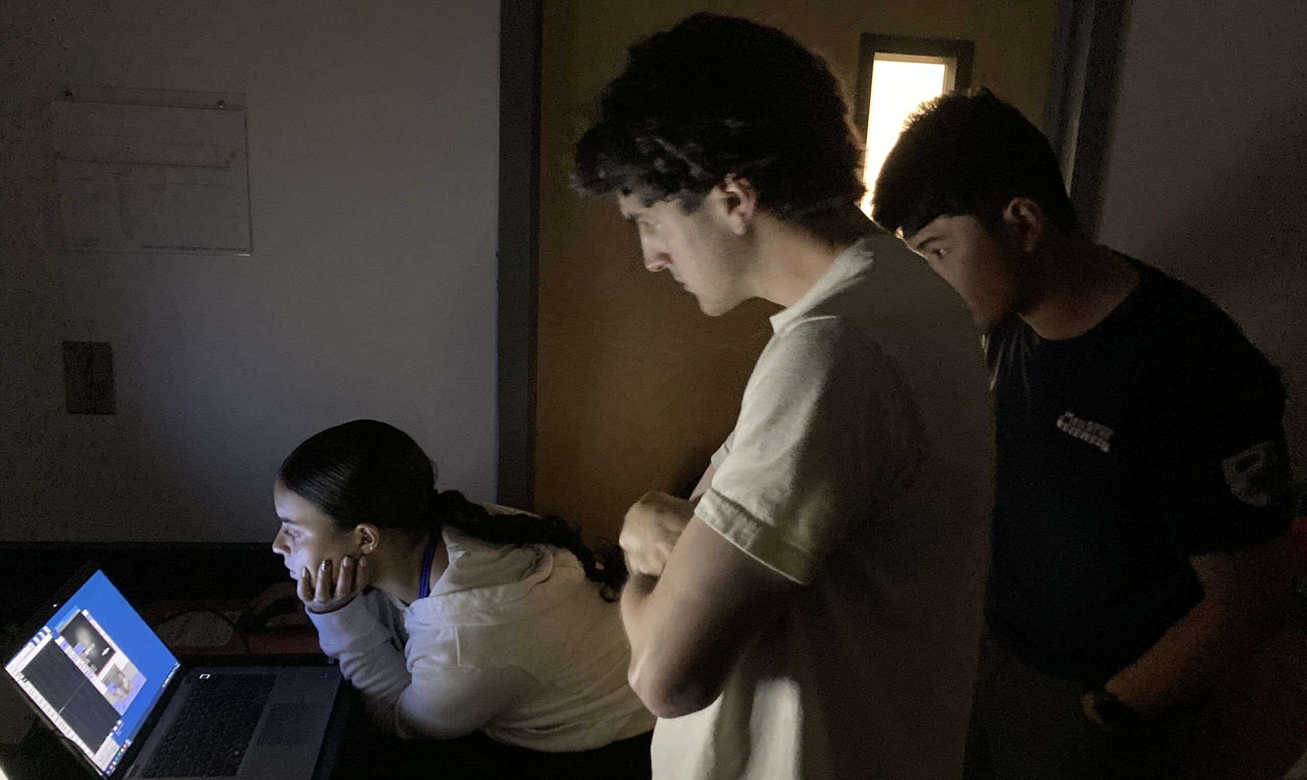 Three people in a dimly lit room look at a laptop screen; one is sitting and the other two are standing behind her.