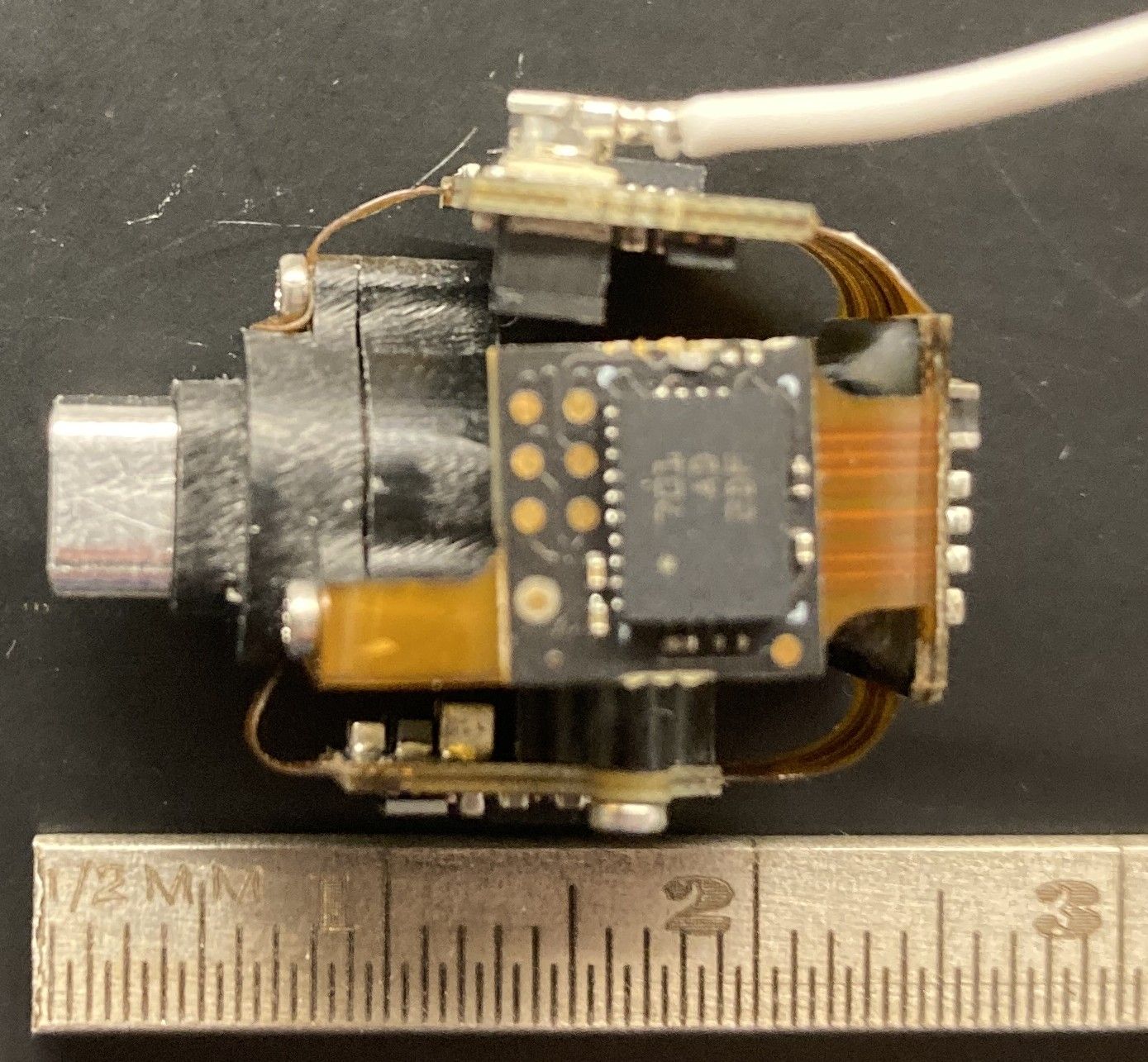 Close-up of a small electronic component with a circuit board and connector, placed next to a ruler showing a length of about 2 centimeters.