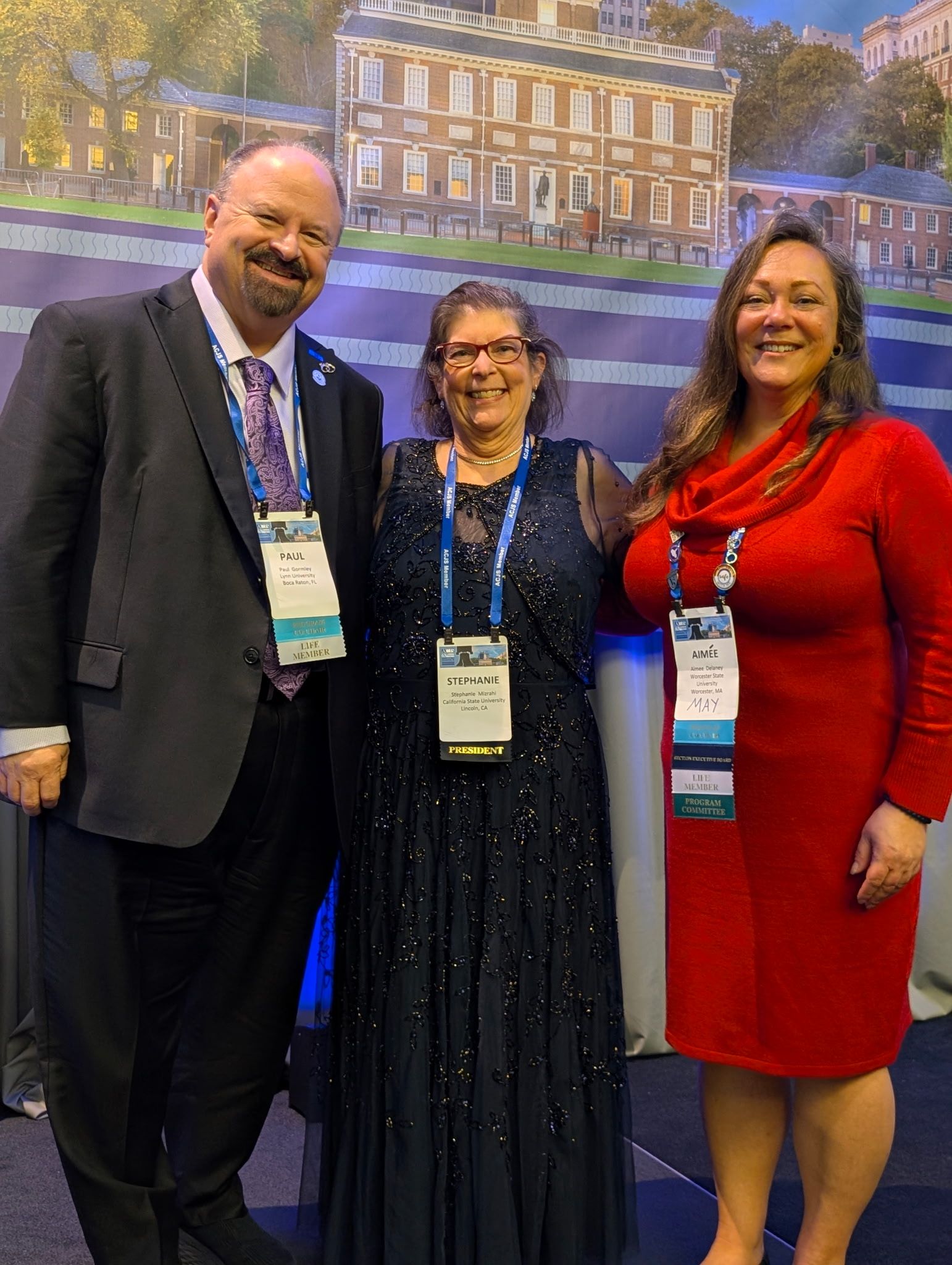 Three people stand together at an event, wearing formal attire and name badges, with a building backdrop behind them.