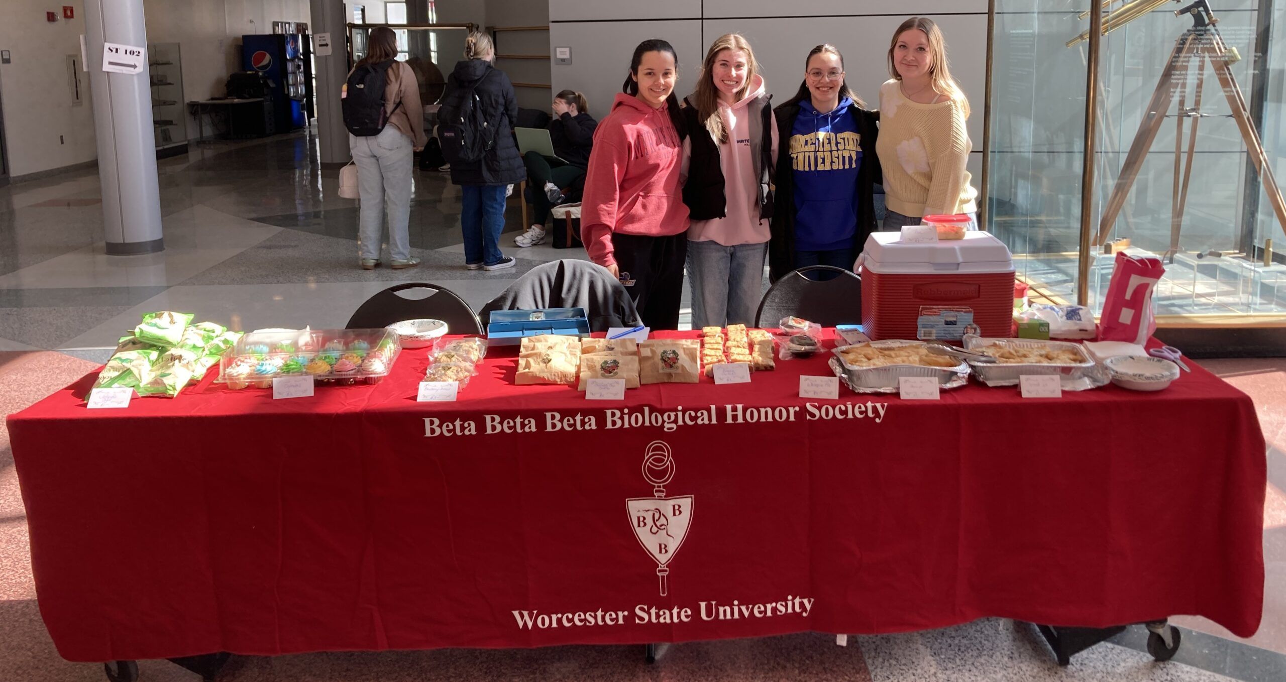 Four people stand behind a table with snacks and baked goods for sale, covered by a red Beta Beta Beta Biological Honor Society tablecloth at Worcester State University.