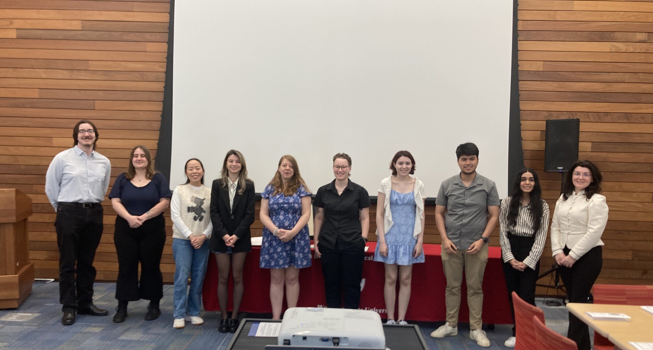 Ten people stand in a row posing for a group photo indoors in front of a projection screen and a wooden wall.