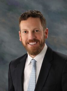 A man with short curly hair and a beard, wearing a dark suit, white shirt, and light blue tie, poses in front of a gray and blue studio background.