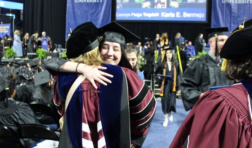 A Worcester State University graduate in cap and gown smiles and hugs a faculty member during a commencement ceremony, with other graduates and attendees in the background.