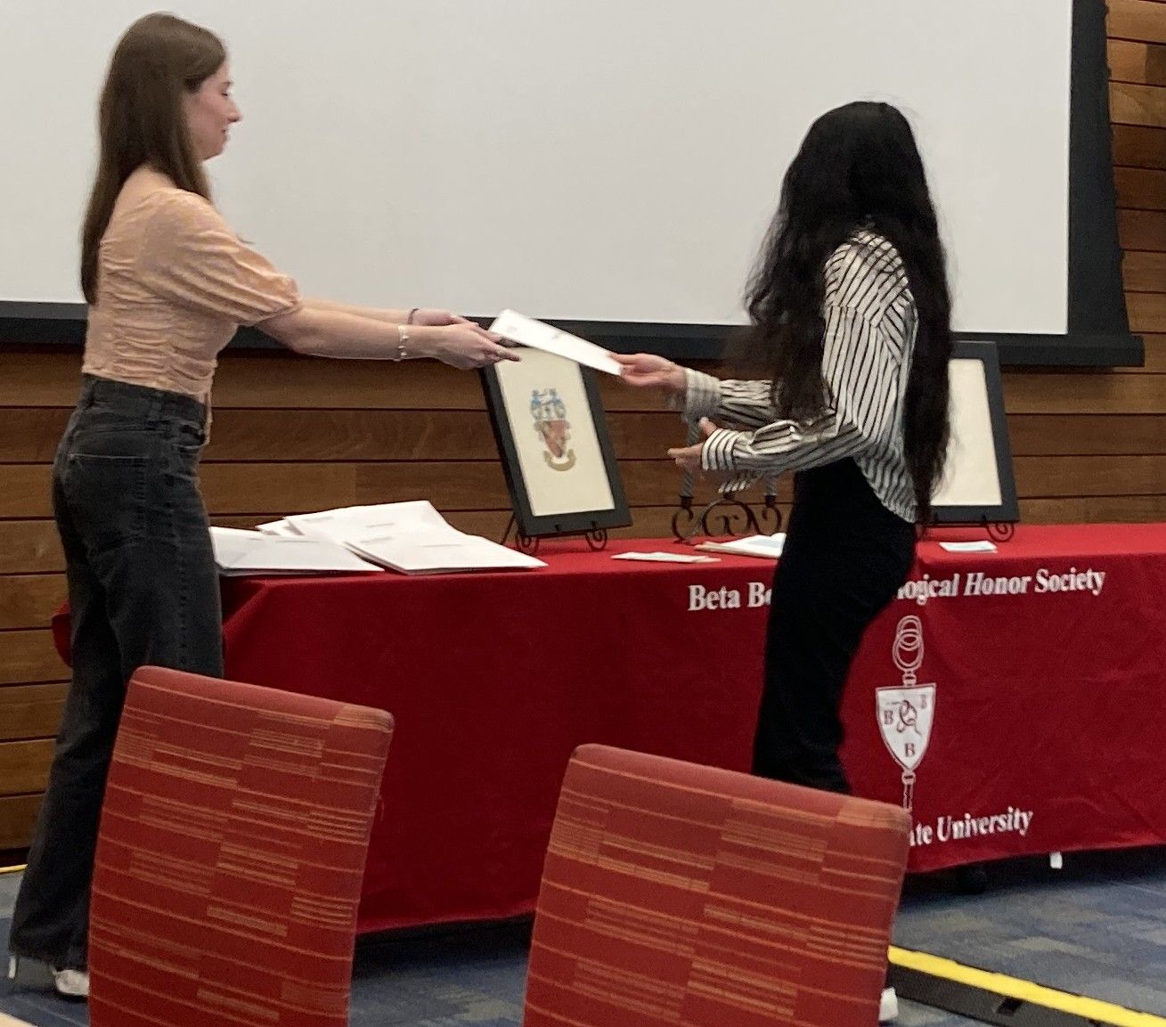 Two women exchange a certificate in front of a table with a red Beta Beta Beta Biological Honor Society cloth and framed images at an event.