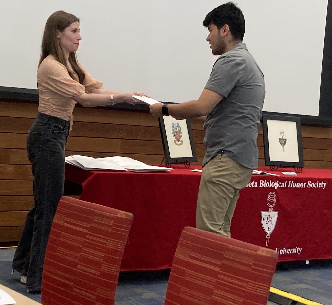 A woman hands a certificate to a man in front of a red Beta Beta Beta Biological Honor Society table with framed items on display.
