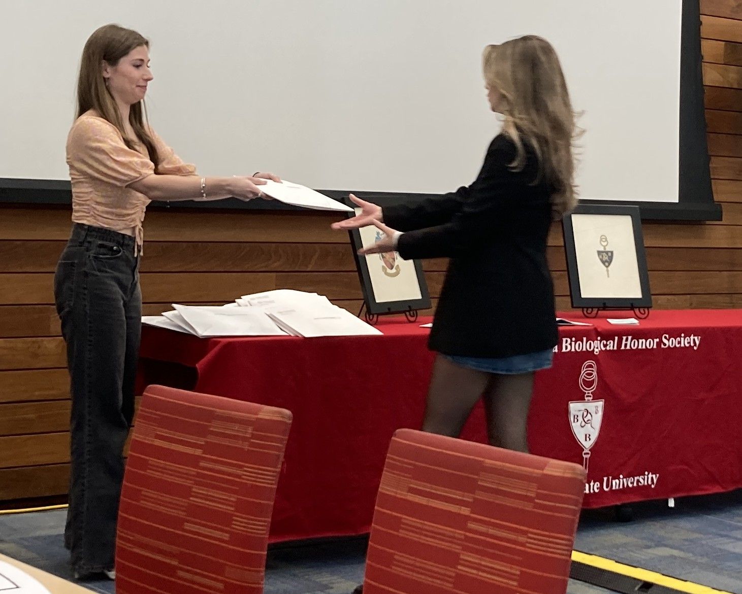 Two women exchange a certificate in front of a red table draped with a Beta Beta Beta Biological Honor Society cloth at an indoor event.