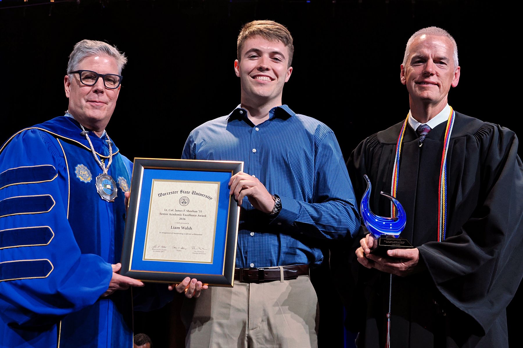 Three men stand together during a formal ceremony, one holding a framed certificate and another showcasing a blue glass award, as they celebrate Academic Achievement Awards.