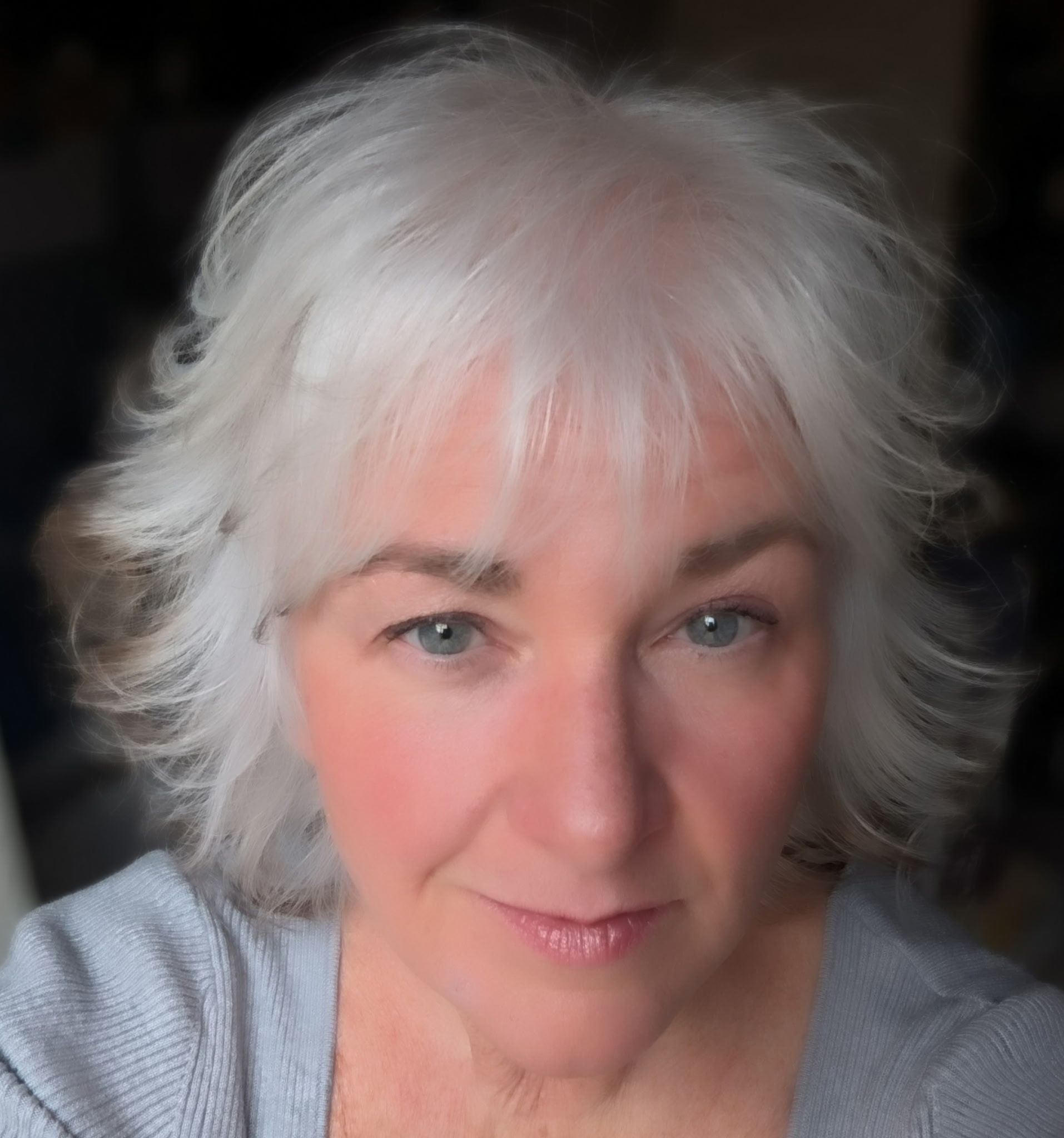 A woman with short, wavy white hair and light skin looks into the camera with a neutral expression, wearing a light gray top.