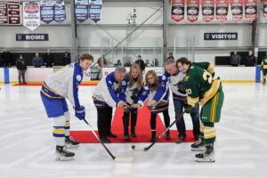 Five adults pose for a ceremonial puck drop on an ice rink, flanked by two hockey players from opposing teams, one in blue and one in green, with banners visible in the background.