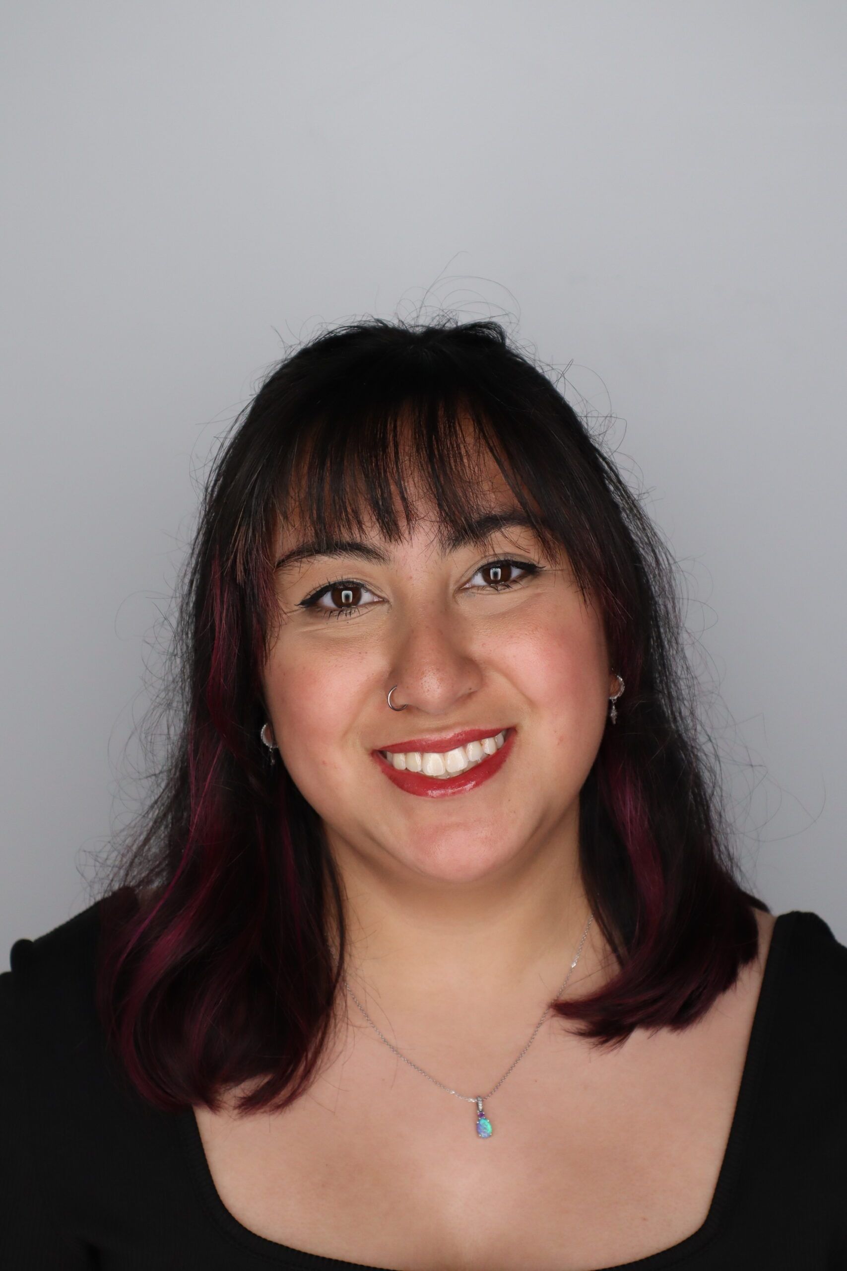 A woman with shoulder-length dark hair and bangs, wearing a black top and a necklace, smiles at the camera against a plain gray background.