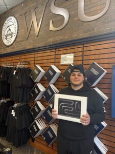 A person in black clothing holds a boxed item while standing in front of a wall display of shirts and books at a Wichita State University store.