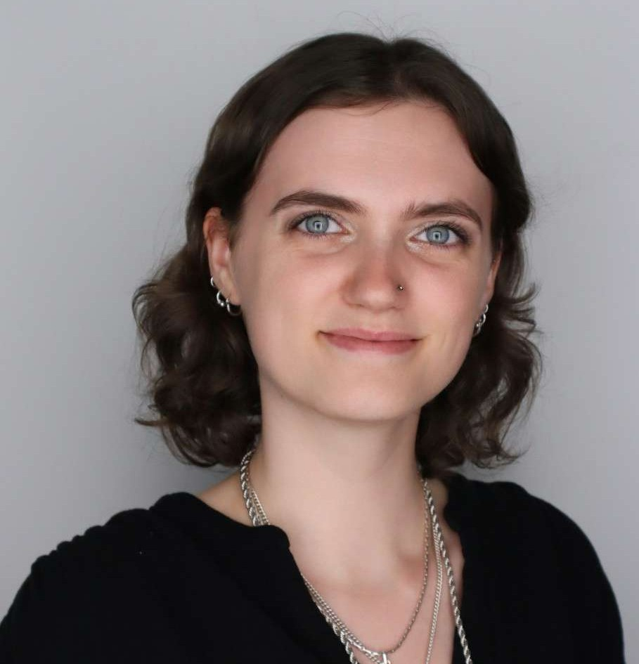 A person with short brown hair and blue eyes, wearing a black top and layered silver necklaces, stands in front of a plain light gray background.