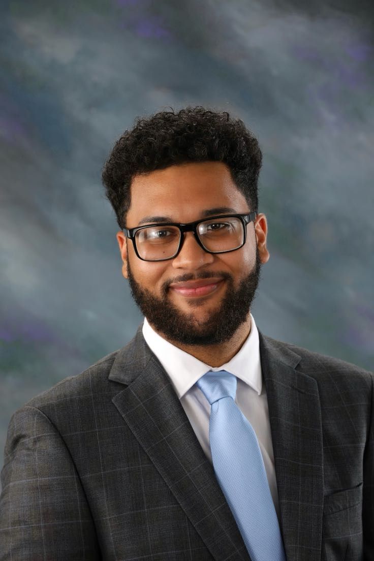 A man with curly hair, glasses, and a beard wears a gray plaid suit, white shirt, and light blue tie, posing in front of a gray and blue studio backdrop.
