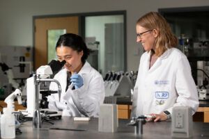 Two scientists in lab coats work in a laboratory; one observes samples under a microscope while the other stands nearby, smiling and watching.