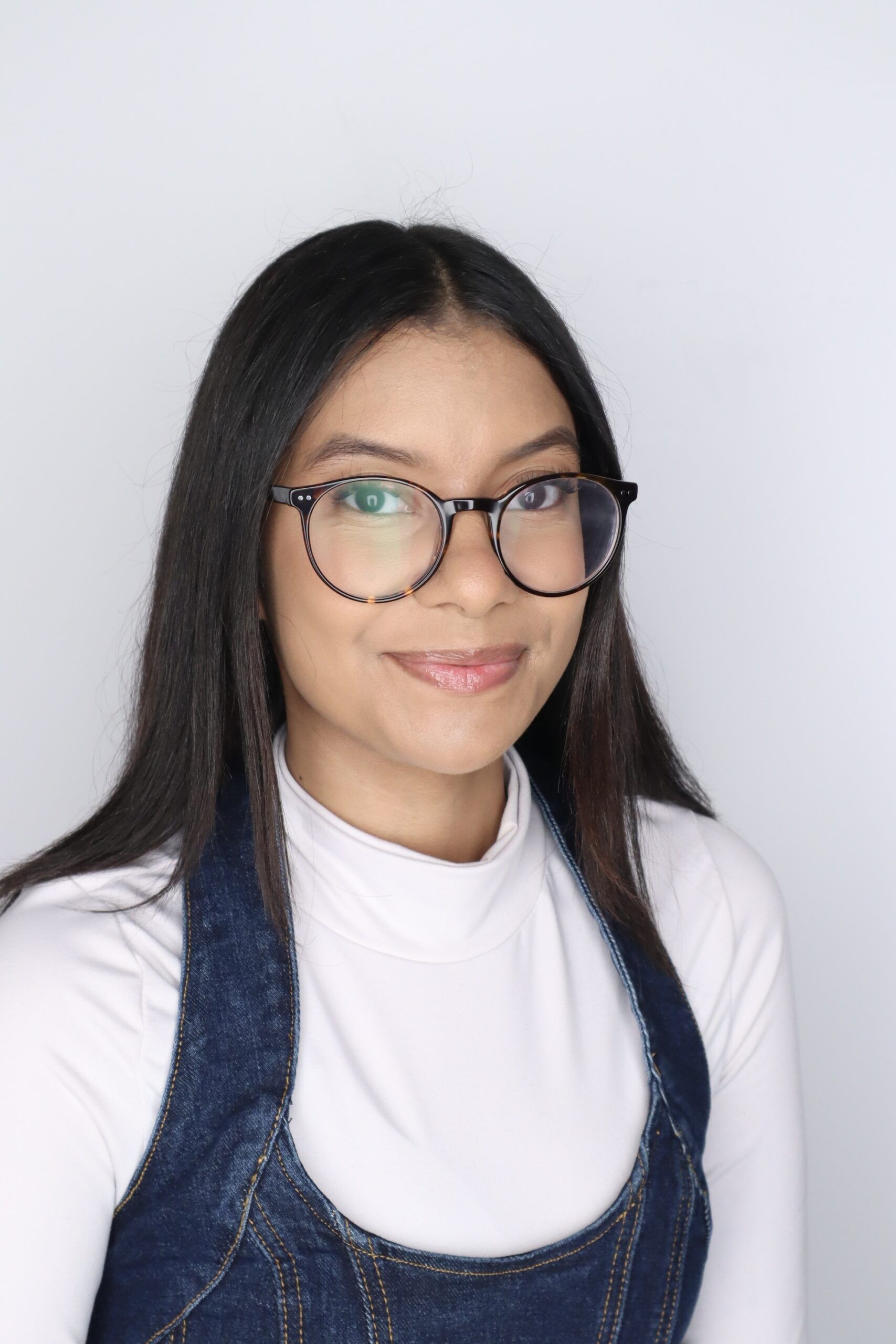 Woman with straight dark hair and glasses wearing a white turtleneck and denim dress, smiling slightly, against a plain light background.