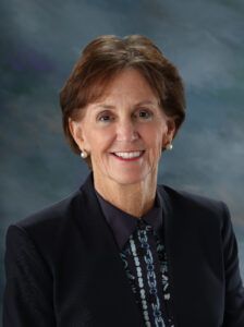 Woman with short brown hair wearing a dark blazer, patterned blouse, and pearl earrings, smiling in front of a blurred blue-gray background.