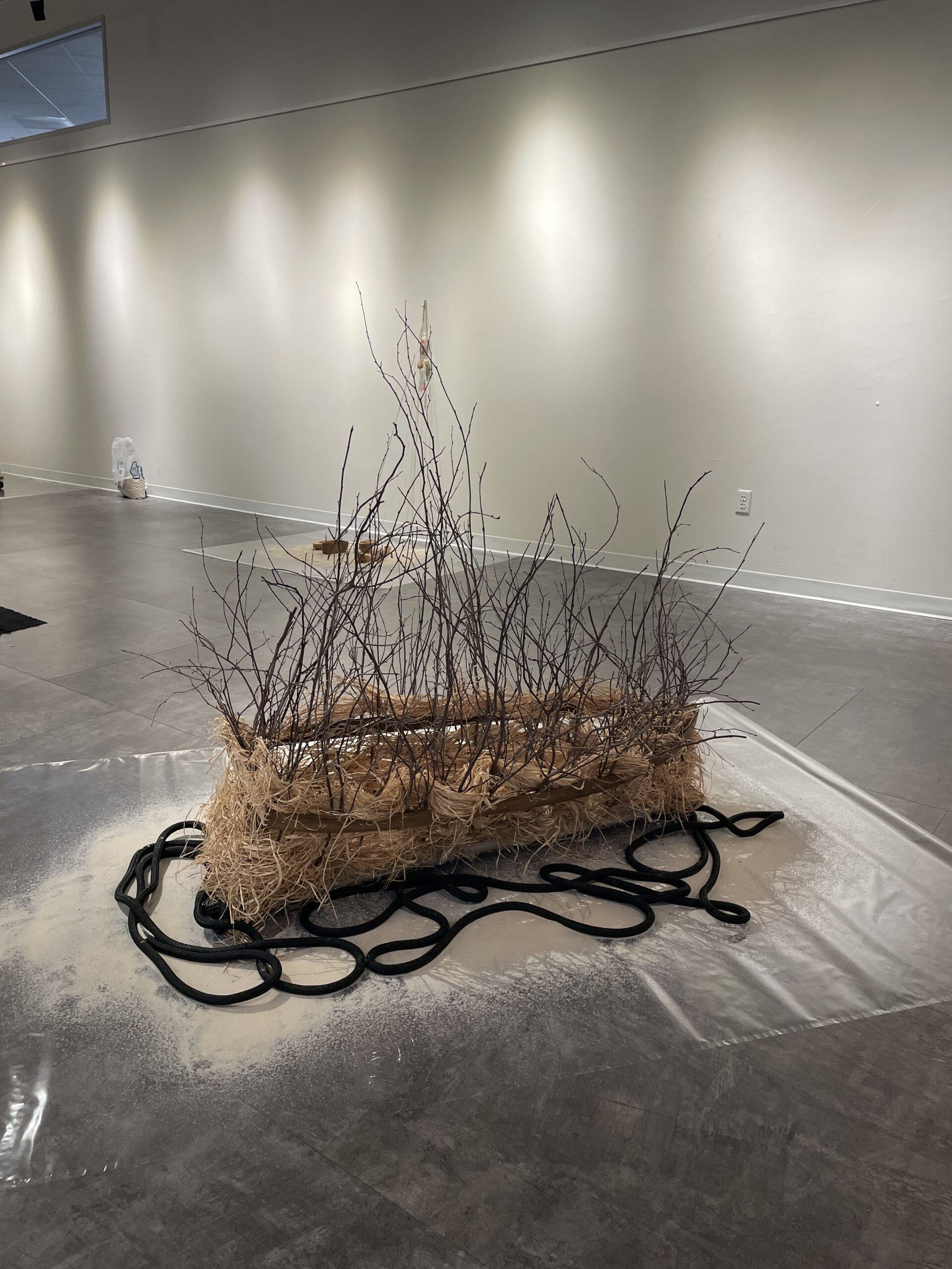 A rectangular bundle of branches and straw rests on black cords, displayed on a plastic sheet in a modern gallery space with neutral walls and floors.
