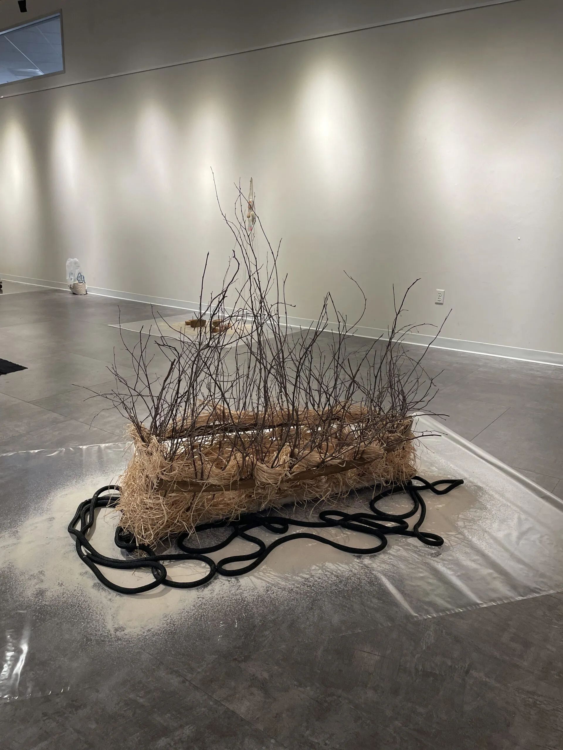 A rectangular bundle of branches and straw rests on black cords, displayed on a plastic sheet in a modern gallery space with neutral walls and floors.