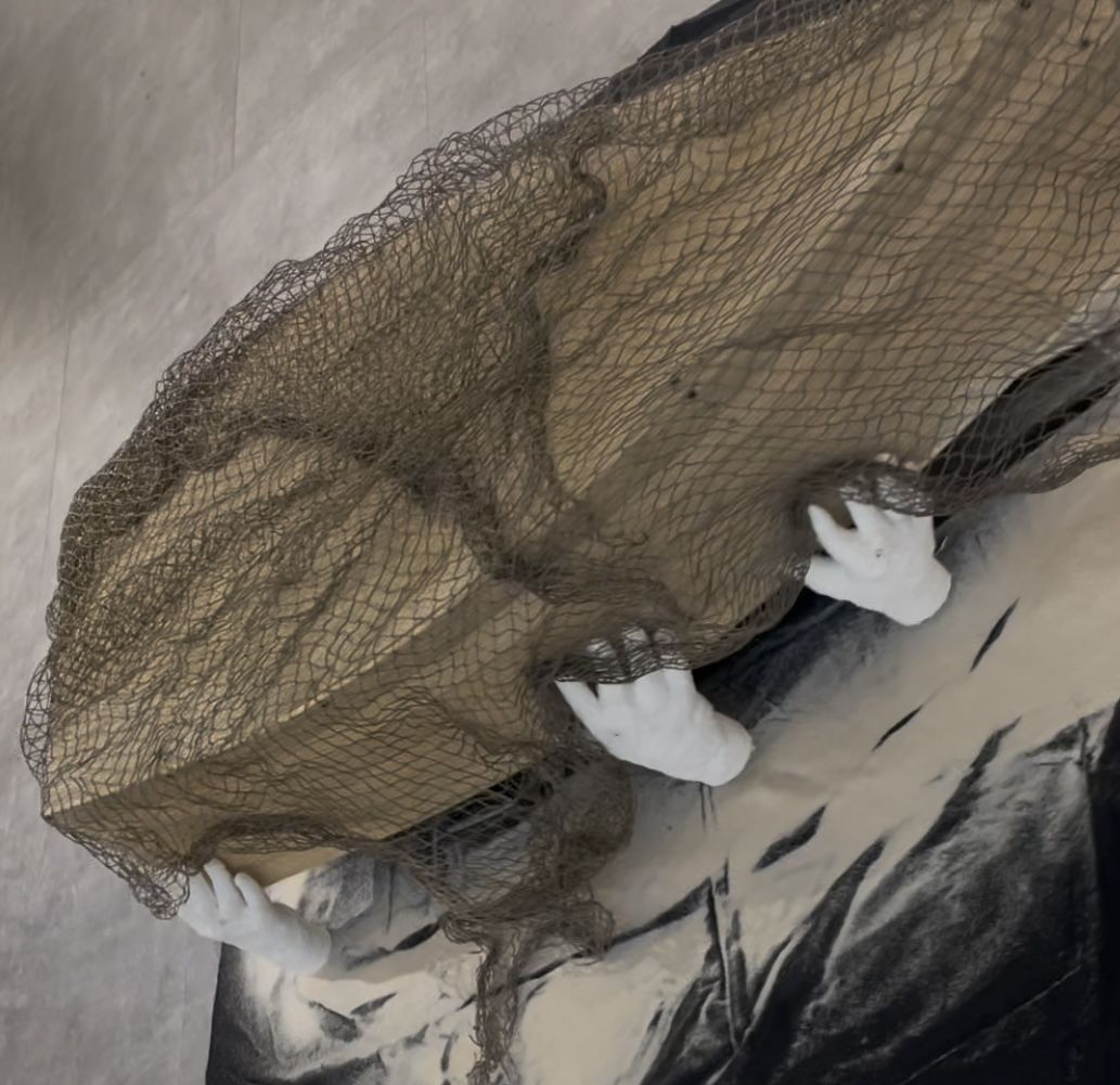 A wooden block covered with mesh is held up by white sculpted hands mounted on a textured gray and black background.