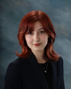 A person with shoulder-length red hair, wearing a black blazer, hoop earrings, and a necklace, poses against a blue-gray studio backdrop.
