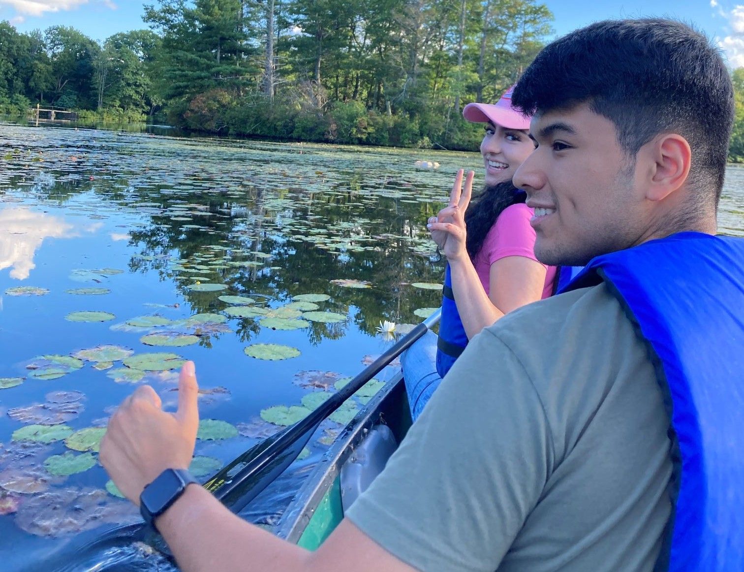 Two people wearing life vests paddle a canoe on a pond with lily pads, smiling and making hand gestures under a blue sky with scattered clouds.