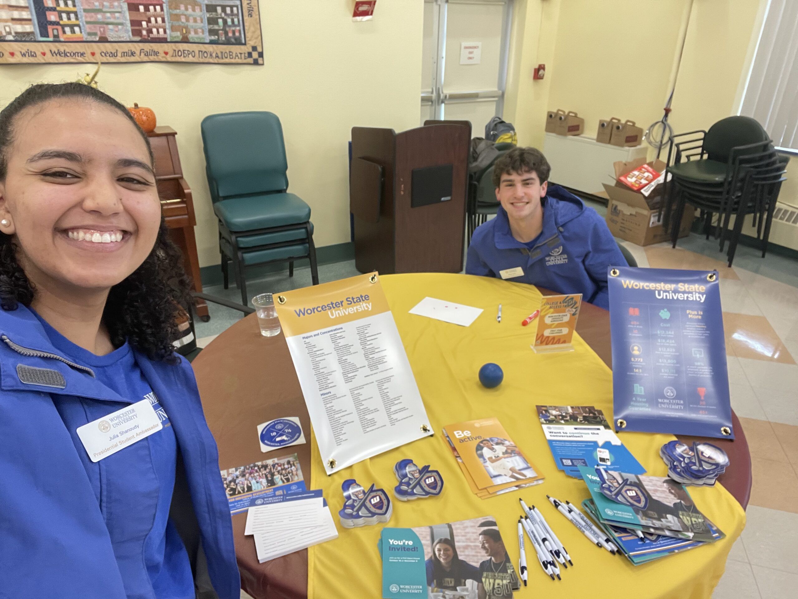 Two people in blue shirts stand by a table displaying Worcester State University materials, including brochures, flyers, pens, and a name list, in a brightly lit room.