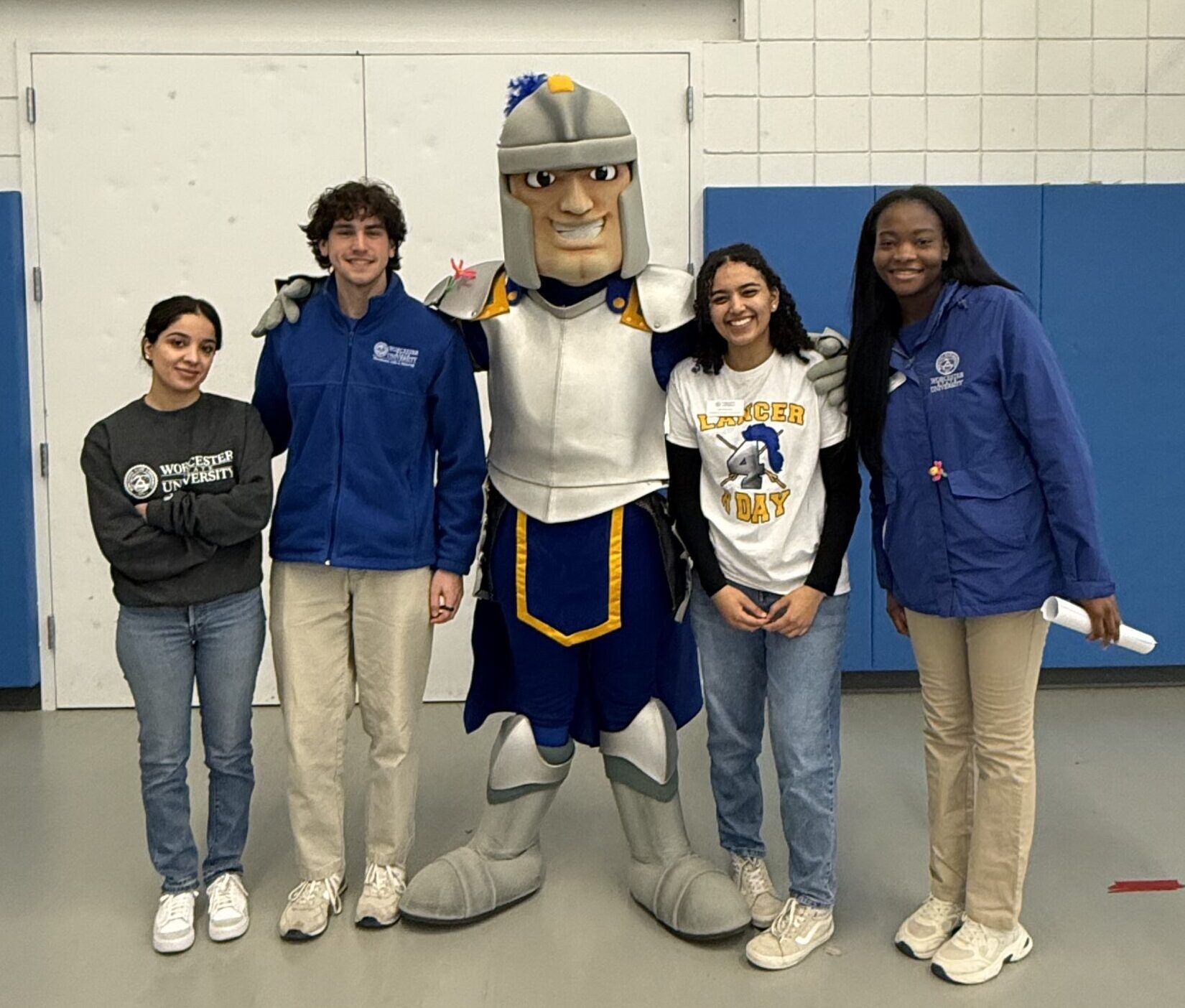 Four students stand indoors posing with a knight mascot. The students are smiling. The setting appears to be a gymnasium with blue and white walls and a gray floor.