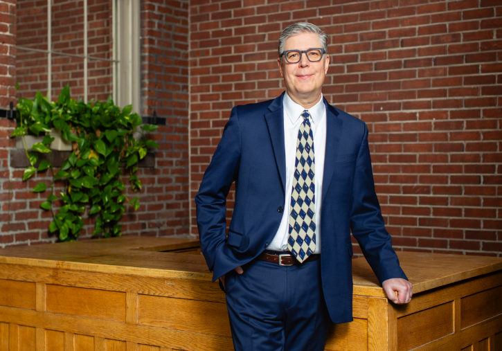 A man in a blue suit and patterned tie stands by a wooden desk in front of a brick wall with a window and a green plant.