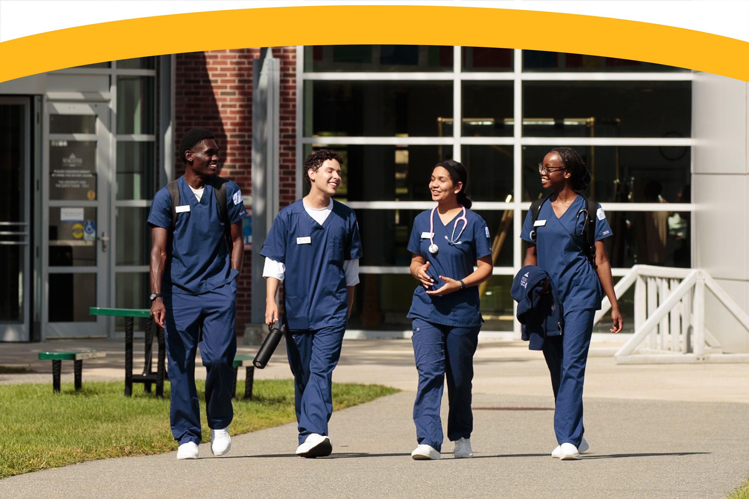 Four nursing students in blue scrubs walk and talk outside a modern building on a sunny day.