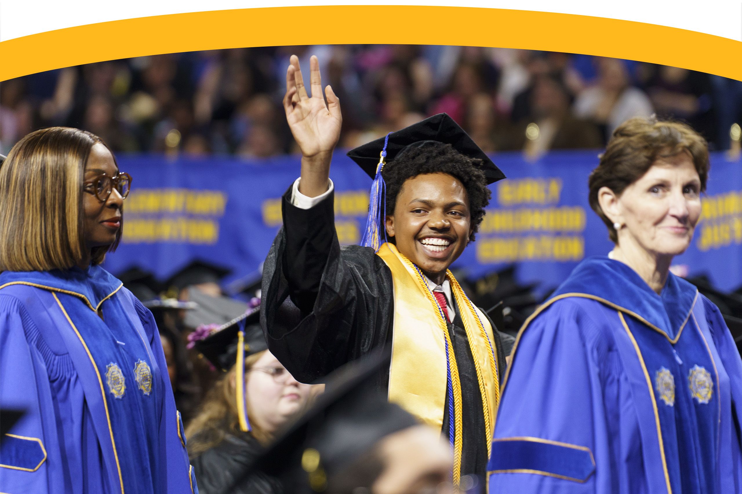 A graduate in a black cap and gown waves and smiles, standing between two faculty members in blue robes at a graduation ceremony.