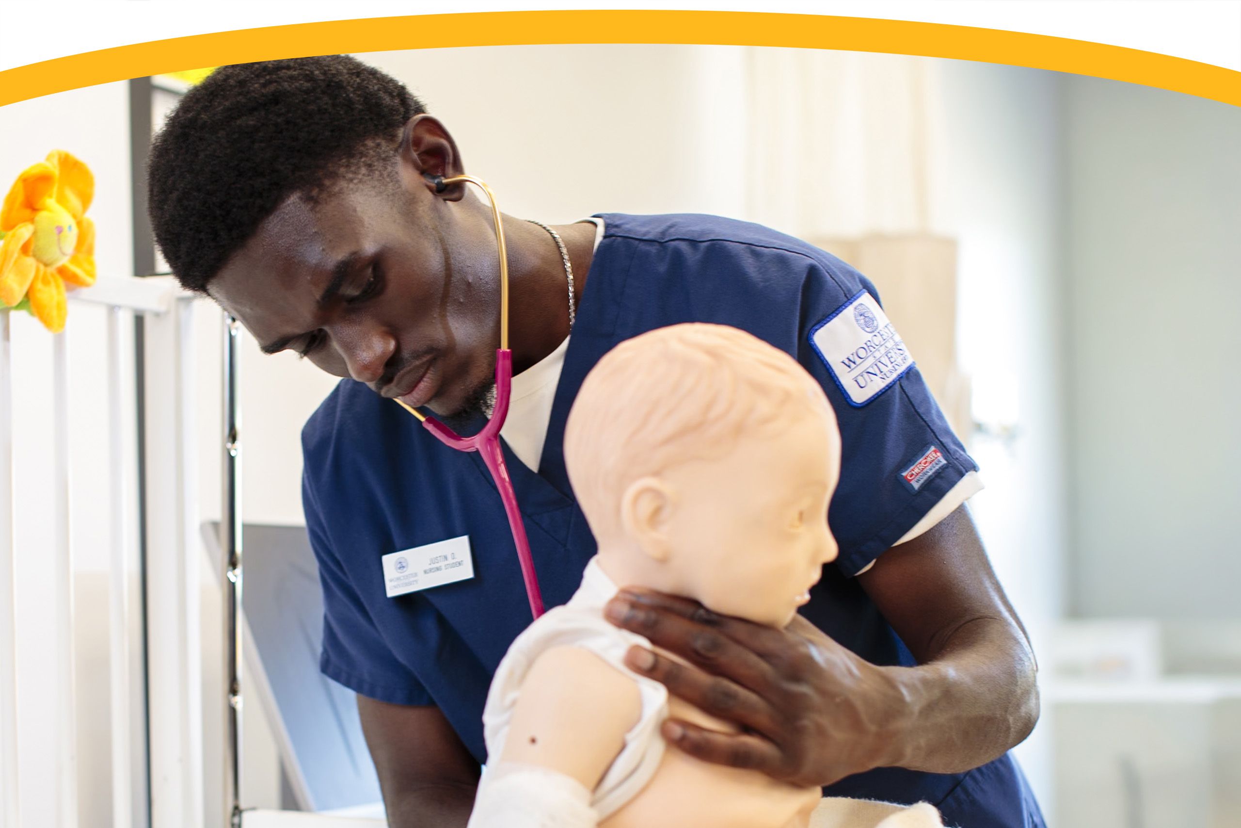 A medical student wearing scrubs uses a stethoscope to examine a child mannequin in a clinical training setting.