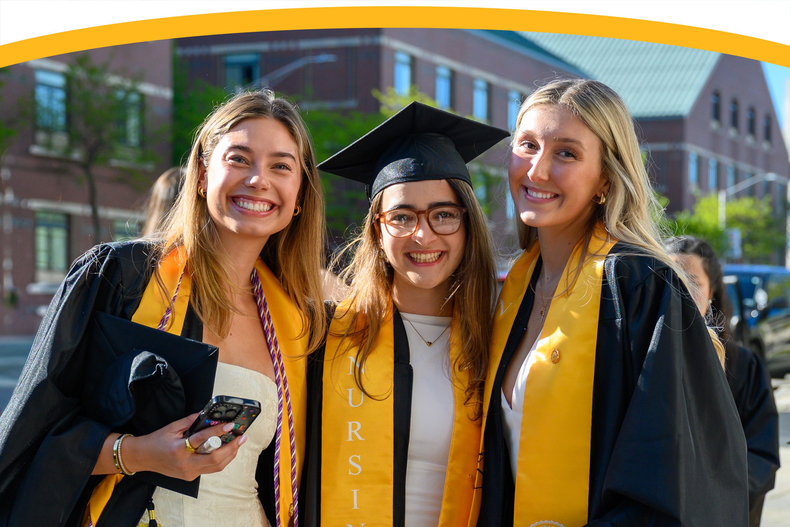 Three young women in graduation caps and gowns smile outdoors, wearing honor cords and stoles, with buildings visible in the background.