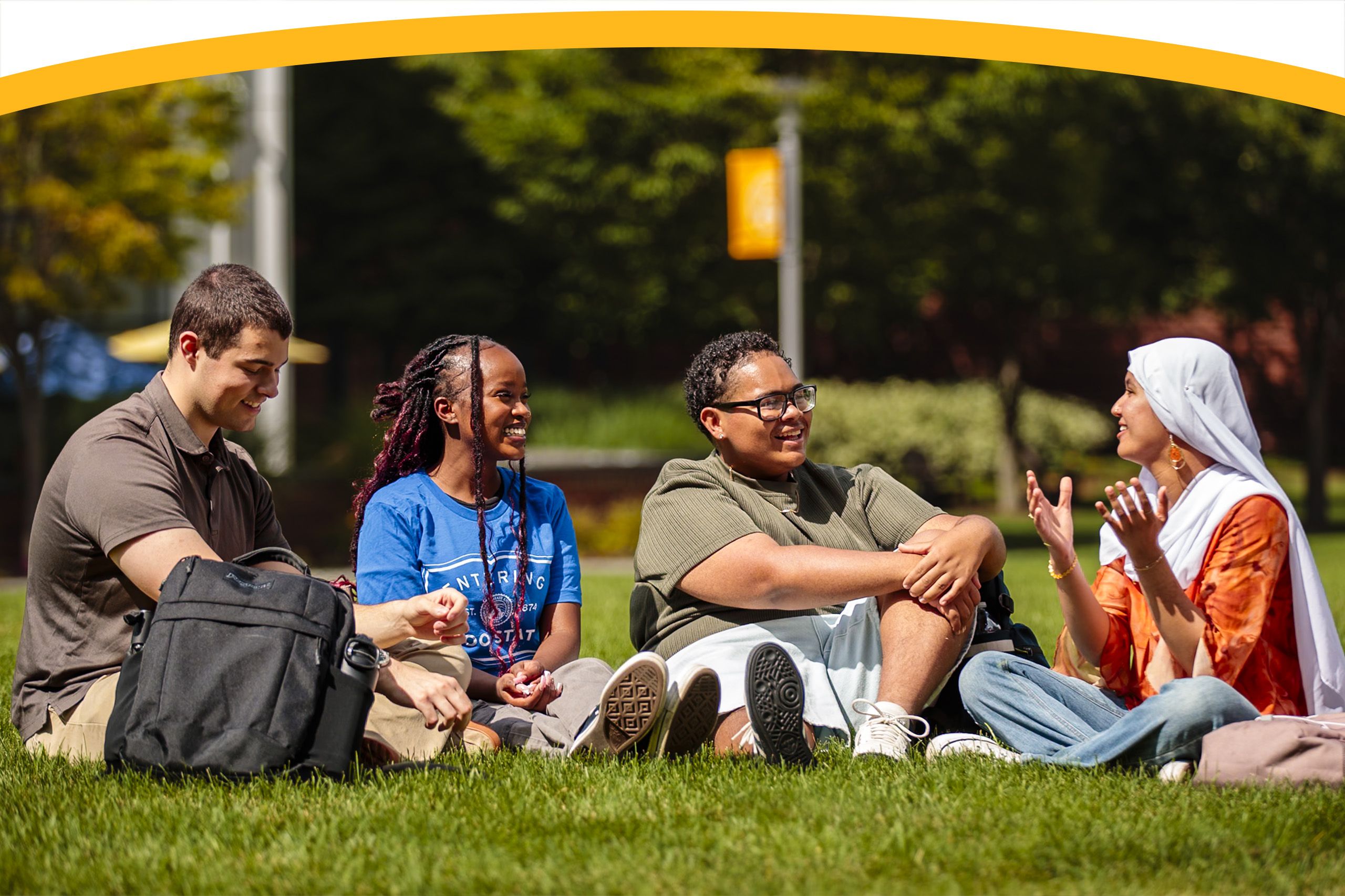 Four students sit on grass outdoors, talking and smiling, with backpacks beside them and trees in the background.