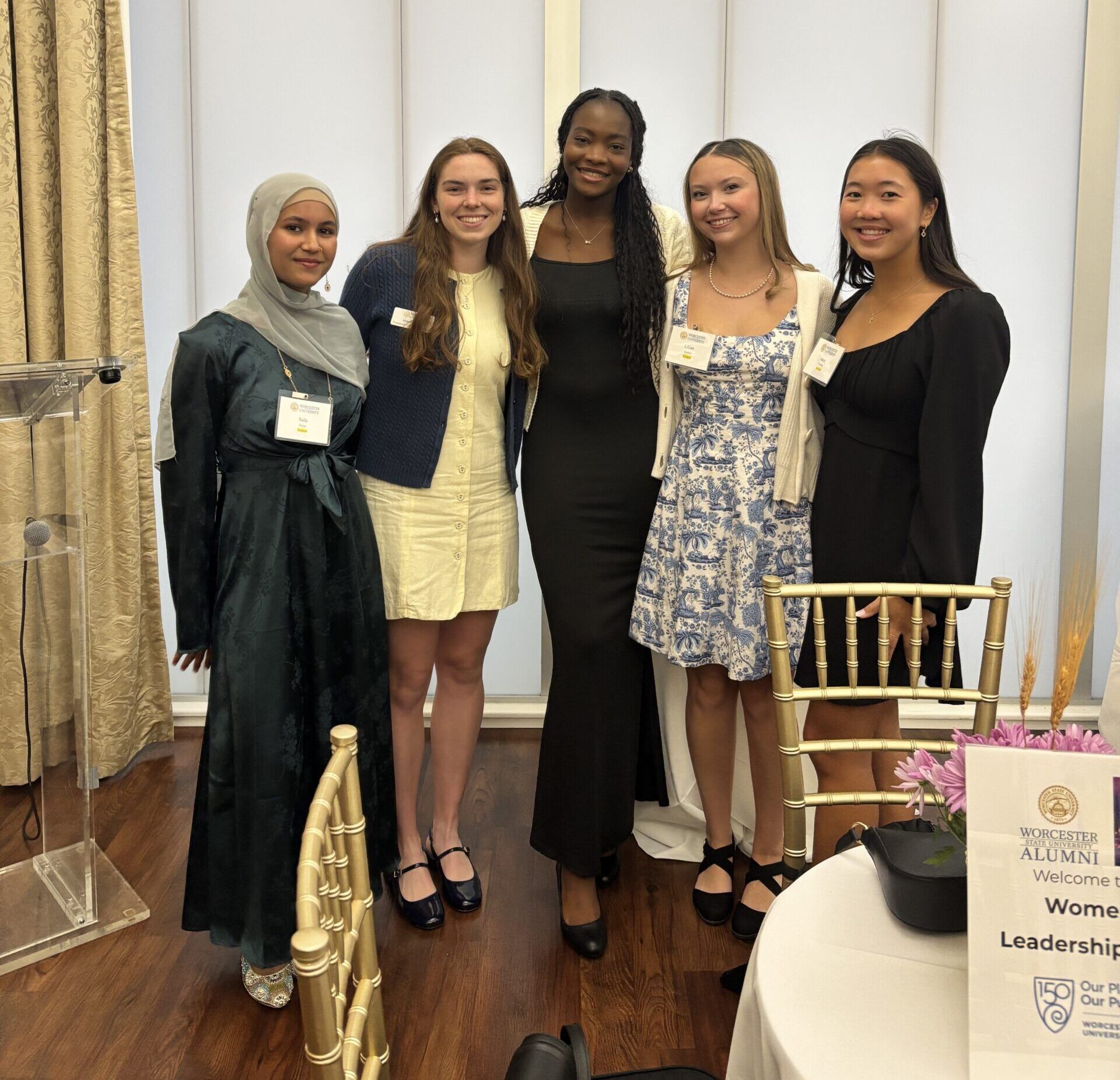 Five women in semi-formal attire stand together and smile for a group photo at an indoor event with gold chairs and floral table decor visible.