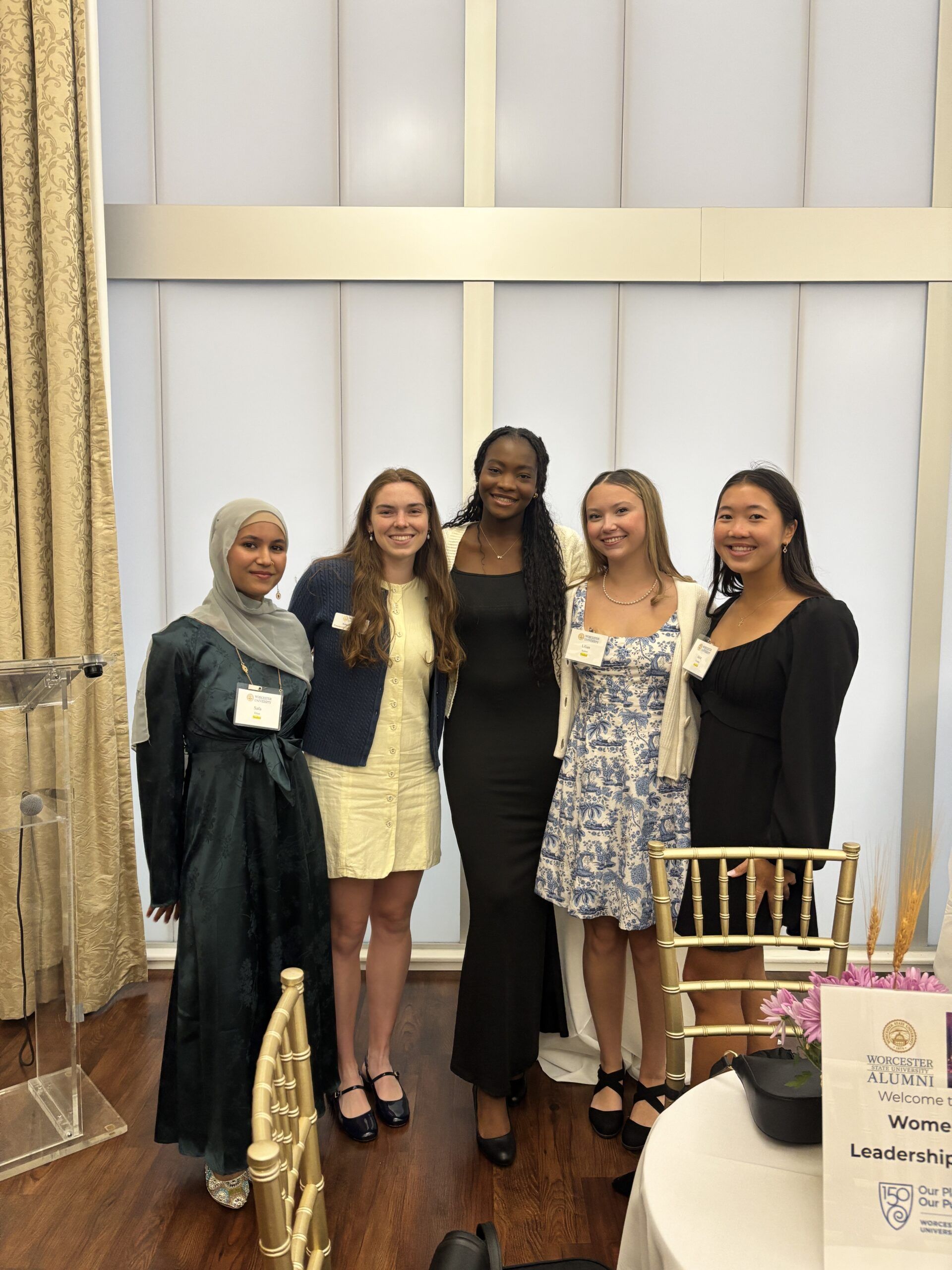 Five women in semi-formal attire stand together and smile for a group photo at an indoor event with gold chairs and floral table decor visible.