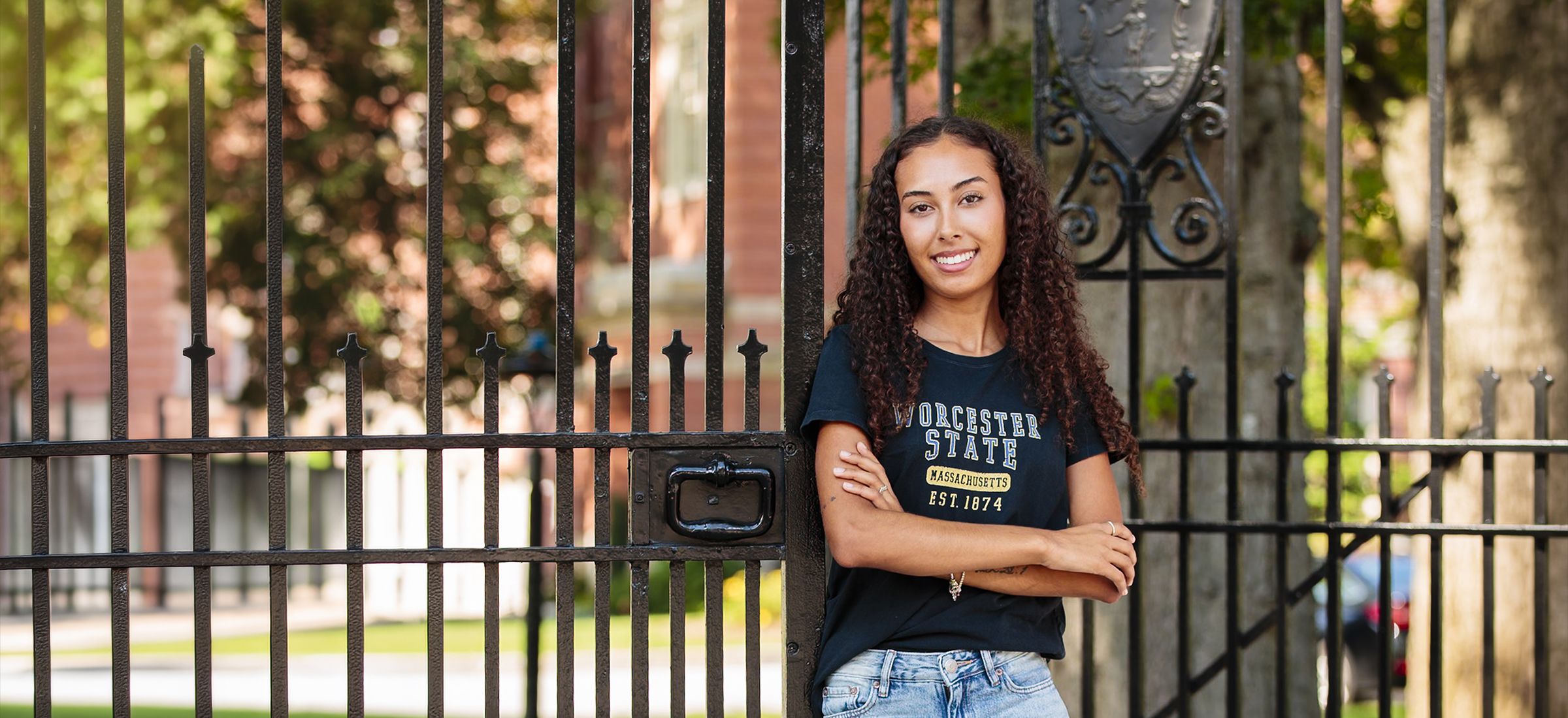 A smiling woman wearing a Worcester State t-shirt stands leaning against an ornate black metal gate.