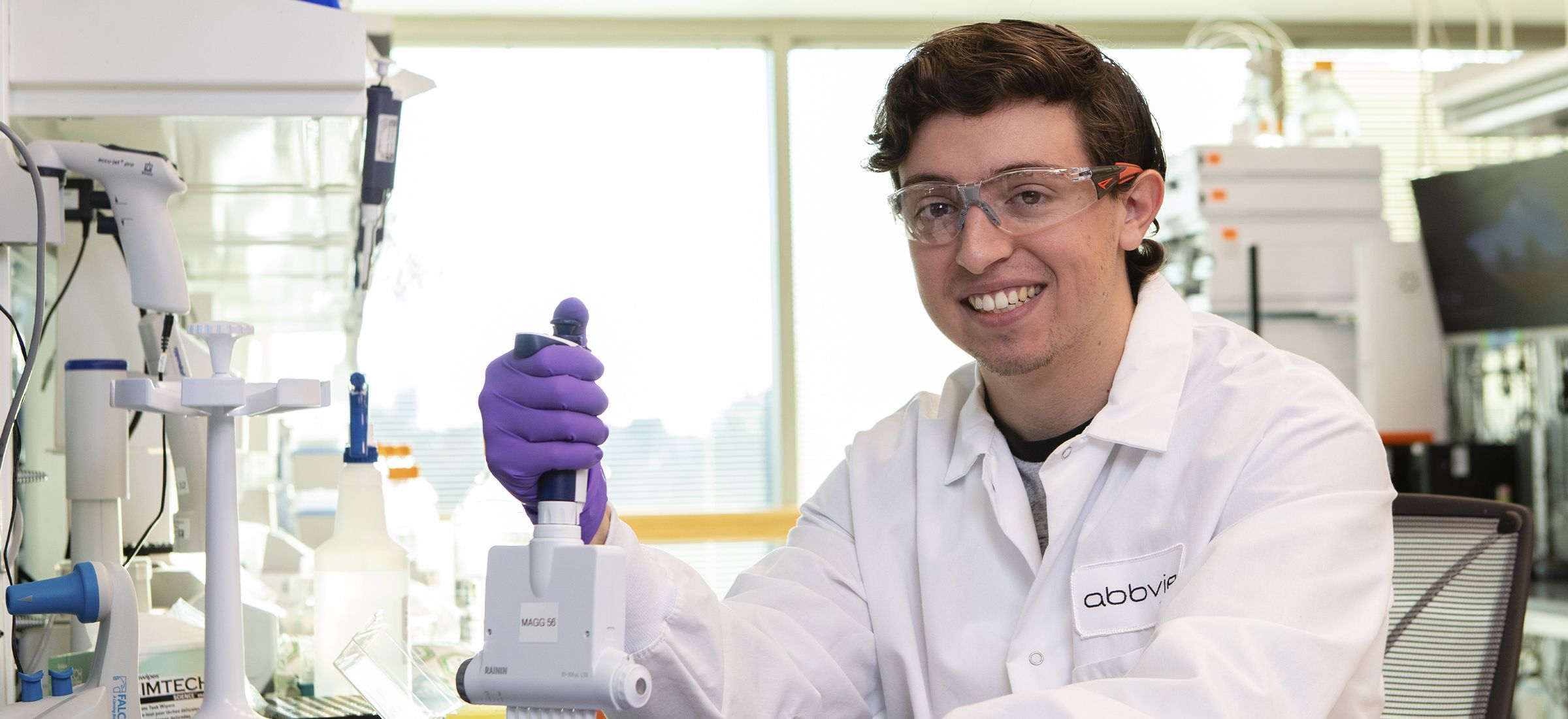 A smiling scientist wearing a white lab coat and safety glasses holds a pipette in a laboratory.