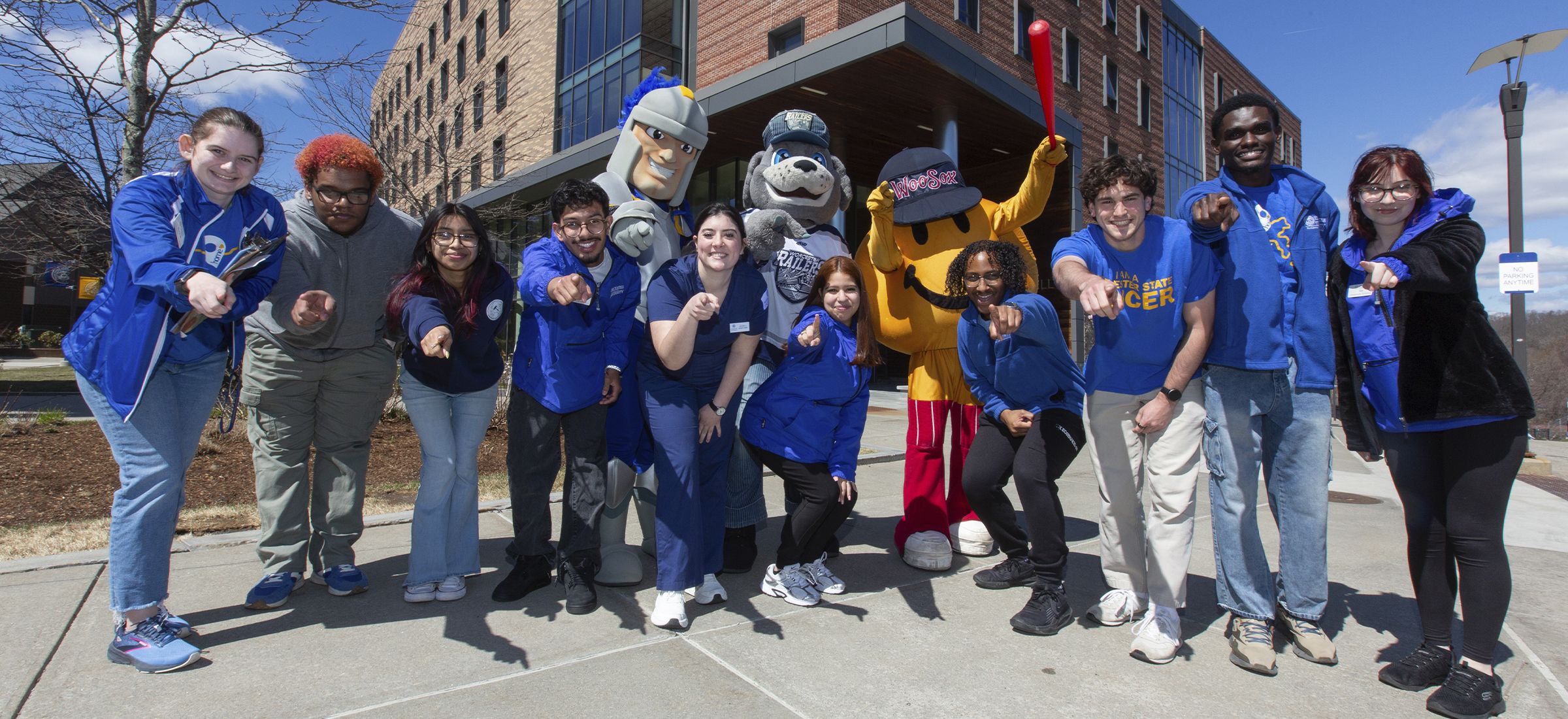 A group of students and university mascots standing outside a brick building, all pointing toward the camera.